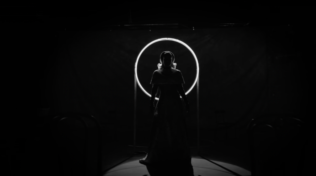 In the 15-second black and white teaser, we see the camera move across the room, stopping at Adele wearing a stunning floral gown accompanied with black gloves and a cape on top, only being highlighted from the ring mirror behind her. 